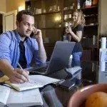 Persona seduta a un tavolo domestico mentre utilizza un computer portatile e prende appunti, con documenti e una tazza sul tavolo; sullo sfondo un’altra persona in cucina tiene in mano una tazza, a rappresentare il contesto domestico del lavoro da remoto.