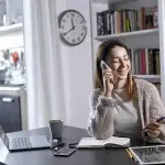 Persona seduta alla scrivania in un ambiente domestico mentre parla al telefono, con un computer portatile aperto, un taccuino e una penna sul tavolo, a rappresentare attività di lavoro agile e gestione professionale da remoto