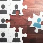 Tessere di puzzle bianche con sagome stilizzate che rappresentano persone, disposte su una superficie in legno; una tessera azzurra con sagoma chiara è separata dal gruppo, a simboleggiare dinamiche di uscita o cambiamento nell’organico aziendale