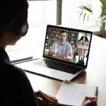 Persona seduta alla scrivania mentre segue una videolezione su un computer portatile, con più partecipanti visibili sullo schermo e un taccuino per prendere appunti, a rappresentare attività di formazione e aggiornamento professionale.