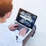 Persona seduta all’aperto mentre utilizza un computer portatile che mostra una schermata con la scritta “Job Search”, a rappresentare attività di ricerca di lavoro e programmi di incentivo all’assunzione.