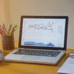 Computer portatile posato su una scrivania che mostra un grafico lineare multicolore, affiancato da un portapenne con matite colorate, un telefono cellulare e documenti, a rappresentare l’analisi dei dati e la gestione della documentazione legata al modello OT23 INAIL.