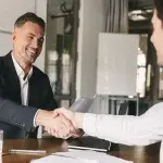 Due persone sedute a un tavolo in un ufficio si stringono la mano sopra documenti e un taccuino, a rappresentare la formalizzazione di un’assunzione o di un accordo professionale.