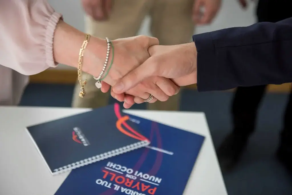 Primo piano di due persone che si stringono la mano sopra un tavolo su cui sono appoggiati documenti rilegati con il logo Fiabilis e grafica aziendale, a rappresentare un accordo professionale o una collaborazione in ambito tecnico amministrativo.