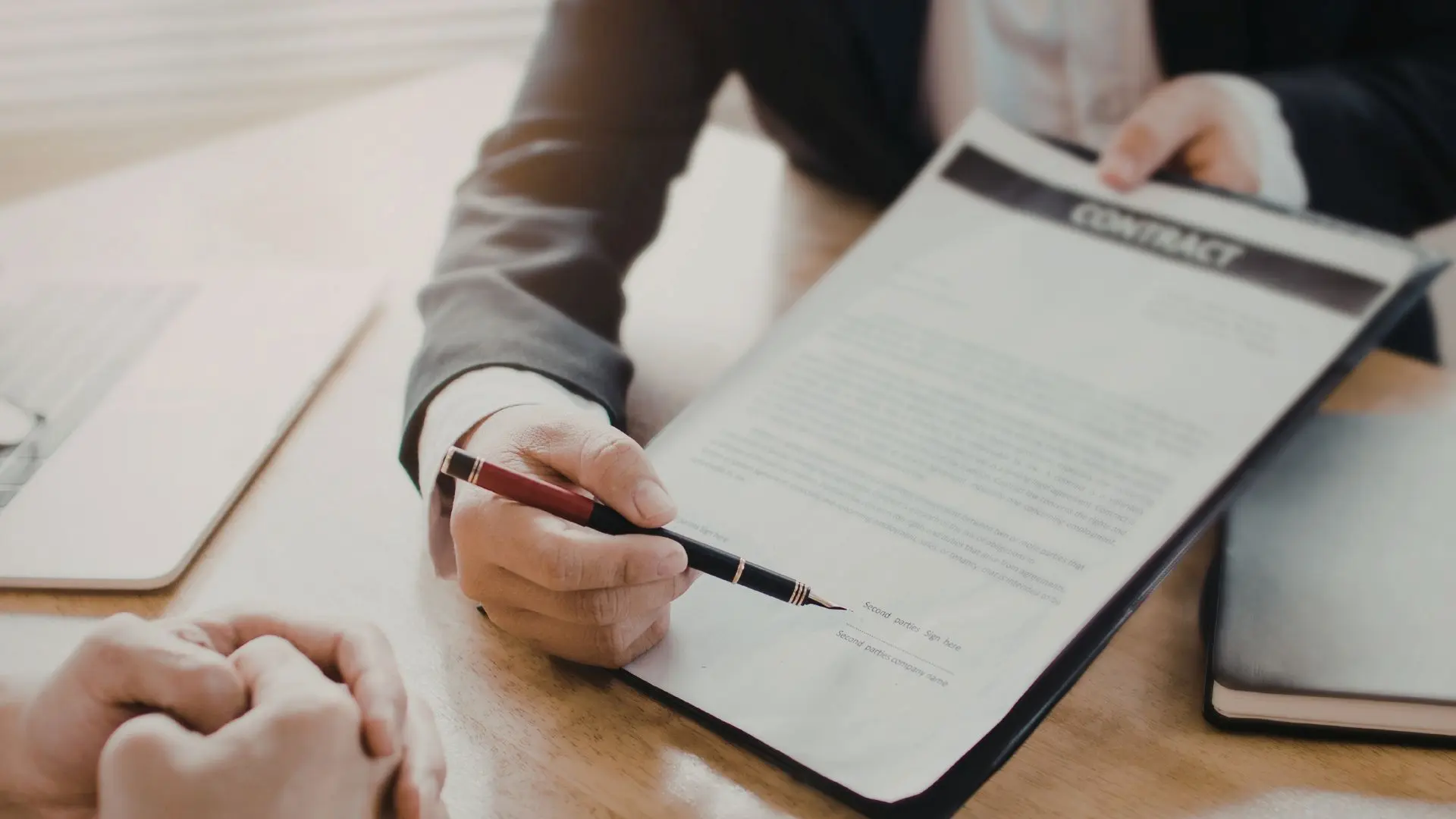 Mano di una persona che indica con una penna un documento contrattuale appoggiato su un tavolo, con un computer portatile e altri materiali da lavoro nelle vicinanze, a rappresentare l’illustrazione e la firma di un contratto di lavoro.