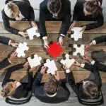 Più persone sedute attorno a un tavolo mentre tengono singoli pezzi di puzzle, con un tassello centrale di colore rosso, a rappresentare la necessità di ricomporre elementi complessi come il calcolo del ticket di licenziamento e la gestione dei contributi.