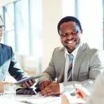 Tre persone sedute attorno a un tavolo in un ufficio luminoso, impegnate in un confronto professionale, con documenti e un tablet sul tavolo, a rappresentare attività di consulenza e gestione degli adempimenti relativi al collocamento mirato.