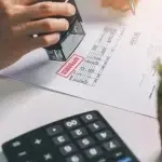 Mano che applica un timbro su un documento amministrativo appoggiato su una scrivania, con una calcolatrice e altri materiali di lavoro, a rappresentare verifiche formali, controlli documentali e procedure di conformità negli appalti