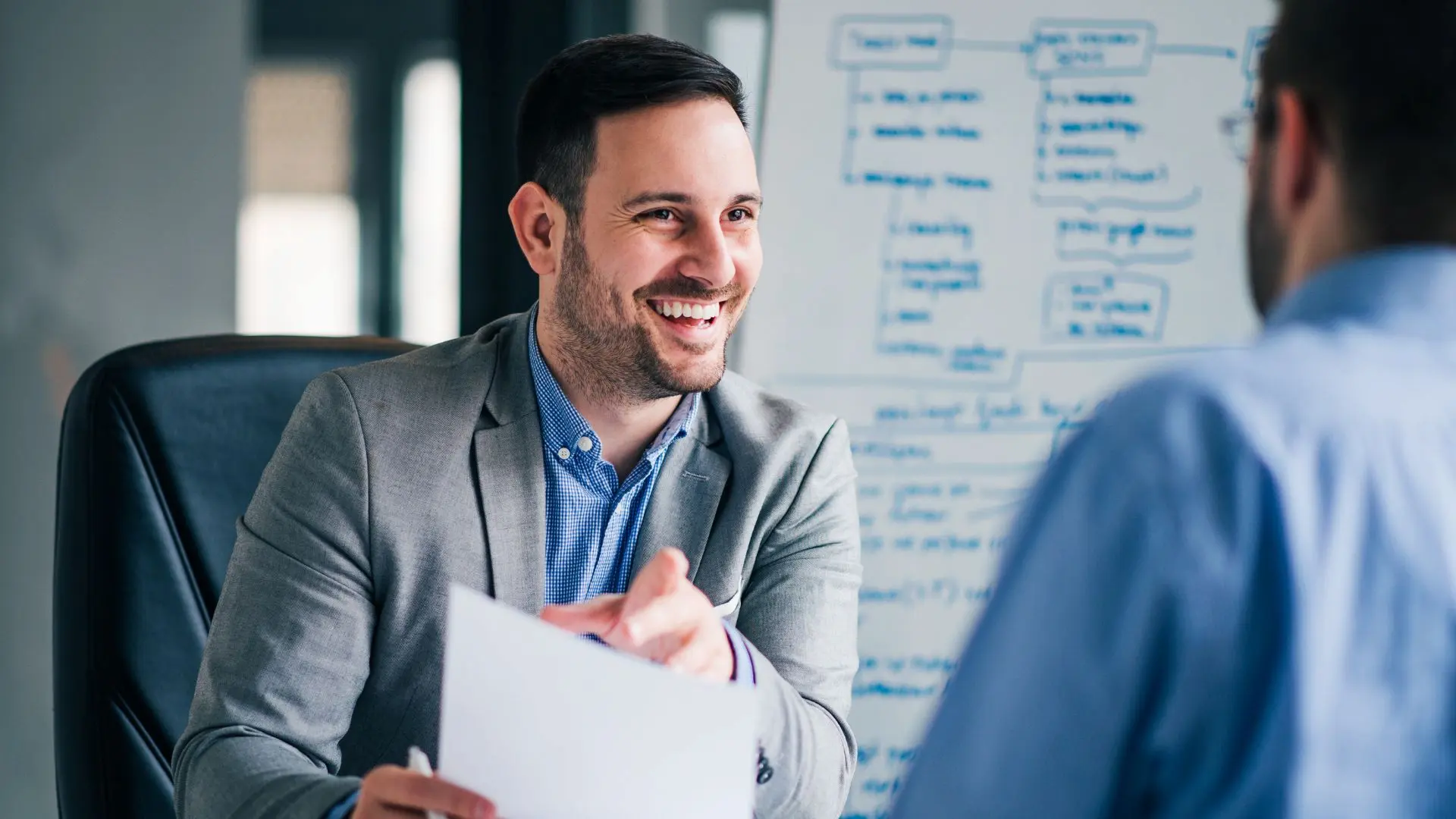 Persona seduta a una scrivania che tiene in mano documenti e dialoga con un’altra persona seduta di fronte, in un ufficio con una lavagna sullo sfondo che contiene schemi e annotazioni, a rappresentare un colloquio o una procedura amministrativa legata alle assunzioni.