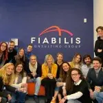 Gruppo di persone riunite in un ambiente d’ufficio davanti a una parete blu con il logo Fiabilis Consulting Group, sedute e in piedi in posa collettiva, a rappresentare spirito di squadra e cultura aziendale