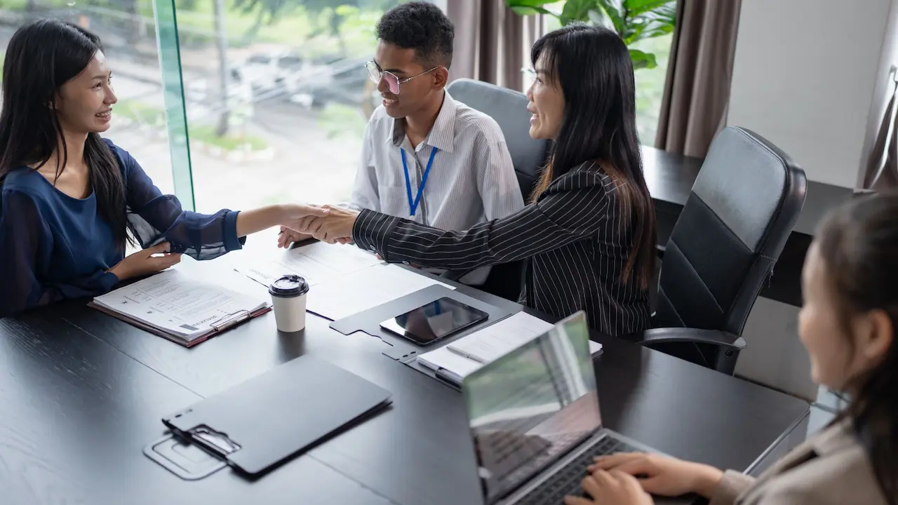Persone sedute attorno a un tavolo in un ufficio mentre due di loro si stringono la mano, con documenti, penne e un computer portatile sul tavolo, a rappresentare procedure di assunzione, gestione dei contratti a termine e attività HR.
