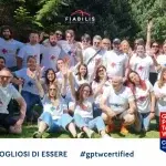 Ampio gruppo di persone riunite all’aperto su un prato, tutte con magliette bianche coordinate, in posa festosa con le braccia alzate; nella grafica compaiono i loghi Fiabilis e Great Place to Work® Certified.