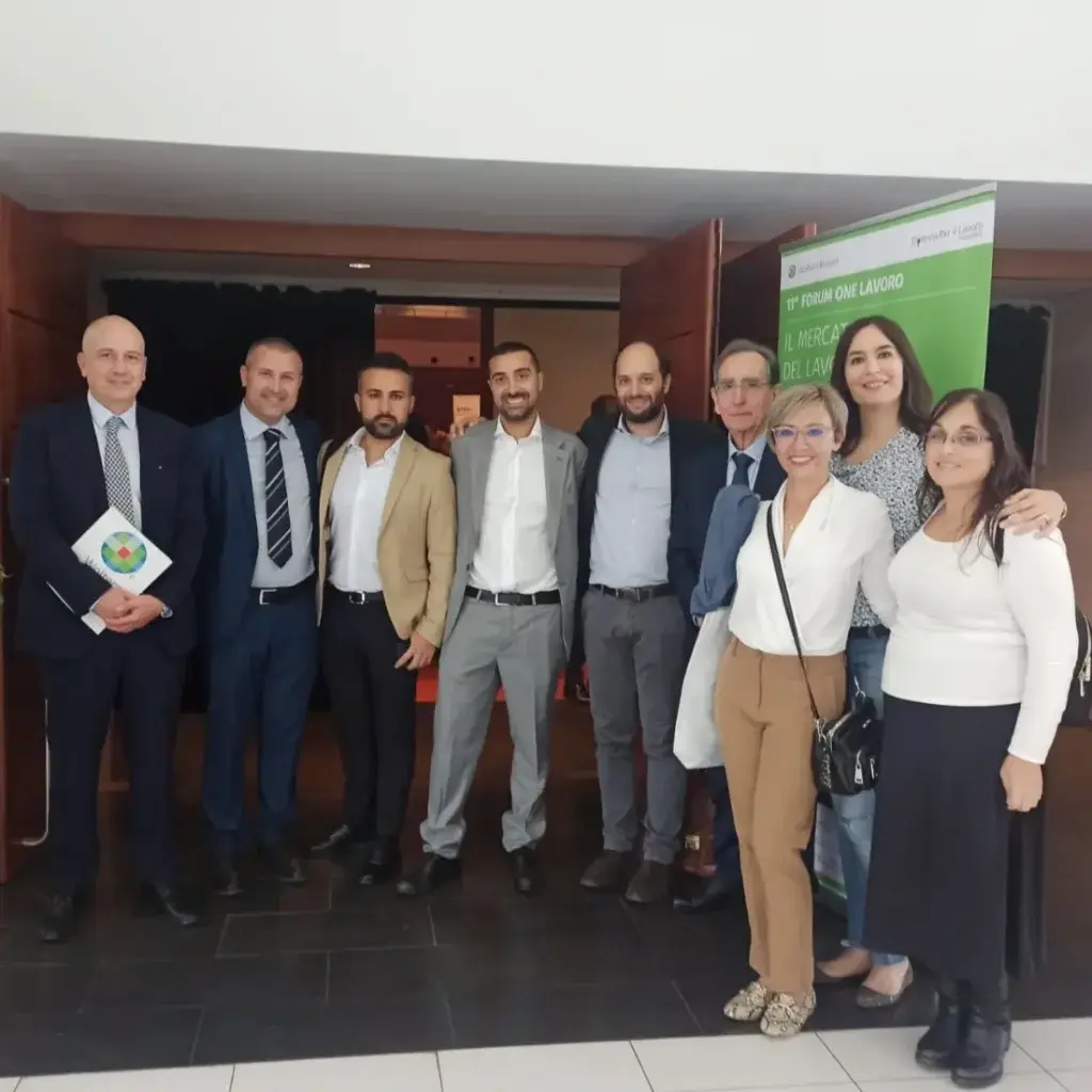 Gruppo di persone in abbigliamento professionale riunite all’ingresso di una sala conferenze, accanto a un roll up verde dedicato all’XI Forum One Lavoro, con sfondo di porte aperte e area evento