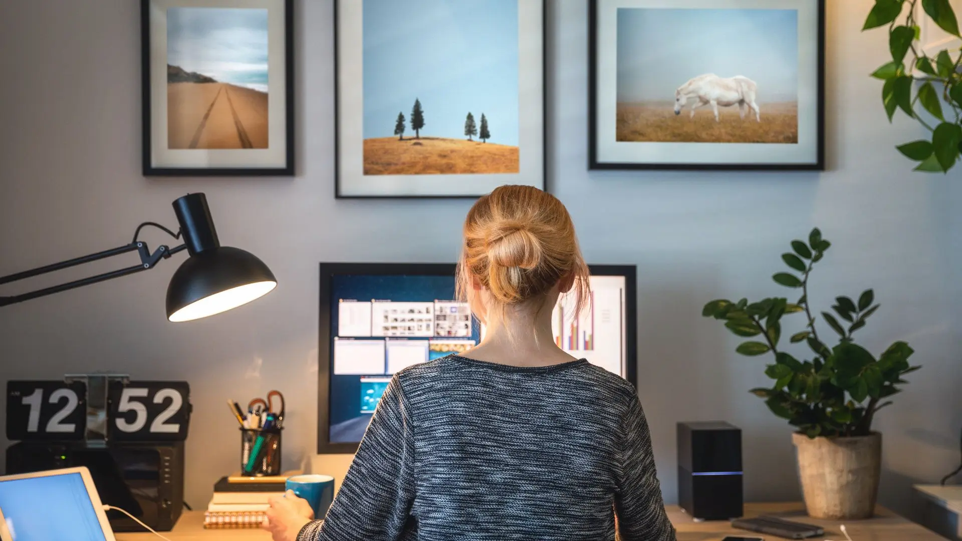 Persona seduta alla scrivania di un ambiente domestico, di fronte a due monitor con documenti e applicazioni aperti, circondata da elementi d’arredo come lampada da tavolo, pianta e quadri appesi, a rappresentare lavoro da remoto e iniziative di welfare aziendale per la conciliazione vita‑lavoro.