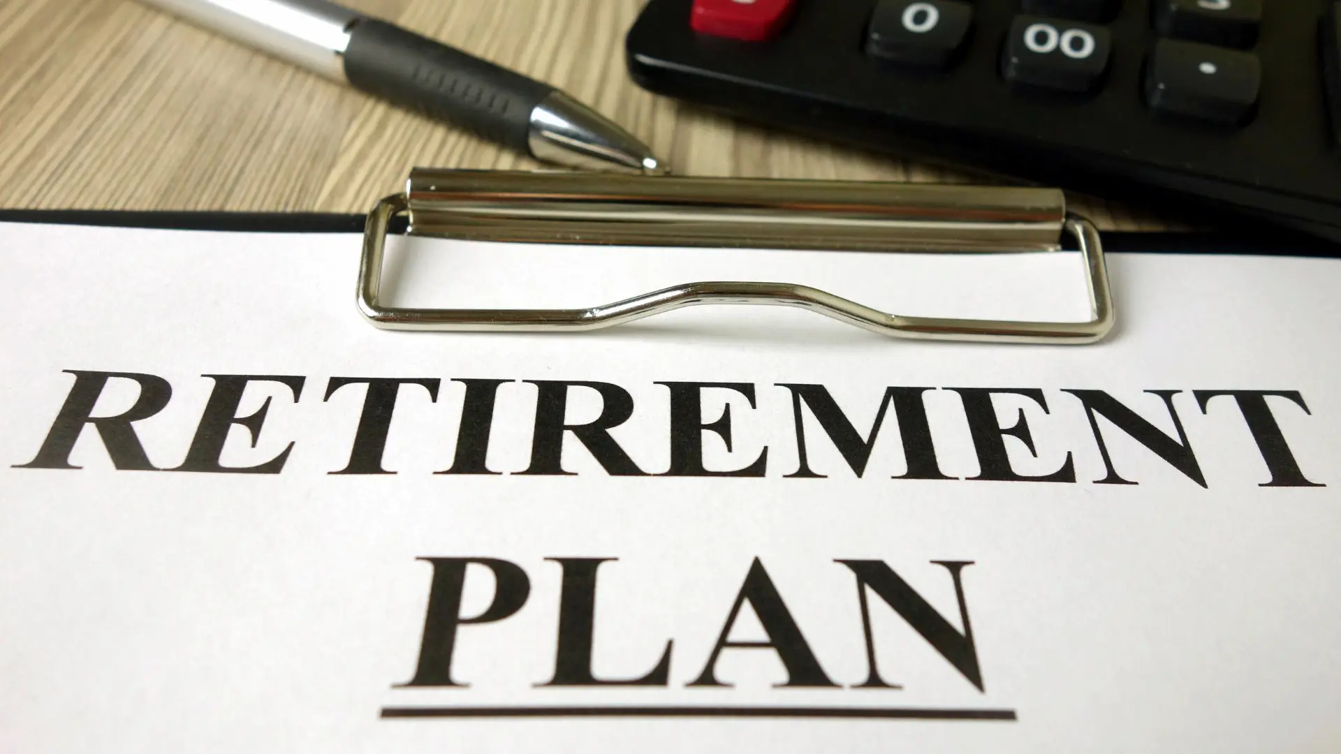Primo piano di un foglio con la scritta “RETIREMENT PLAN” posato su una clip board, con una penna metallica e parte di una calcolatrice sullo sfondo, a rappresentare strumenti e documentazione relativi alla pianificazione della pensione.