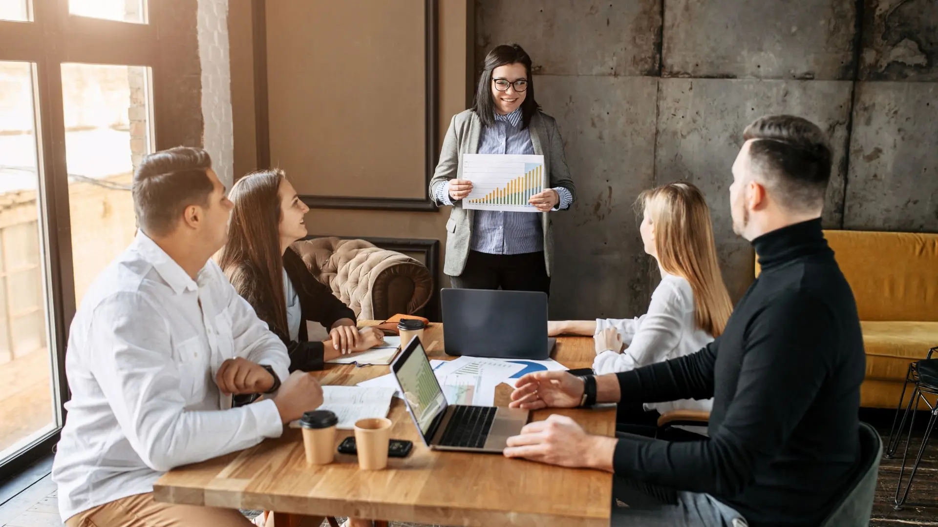 Persone riunite attorno a un tavolo in un ufficio, con laptop, documenti e grafici, mentre una persona in piedi mostra un foglio con un grafico a barre, a rappresentare un confronto tecnico su incentivi all’occupazione e analisi dei contributi.