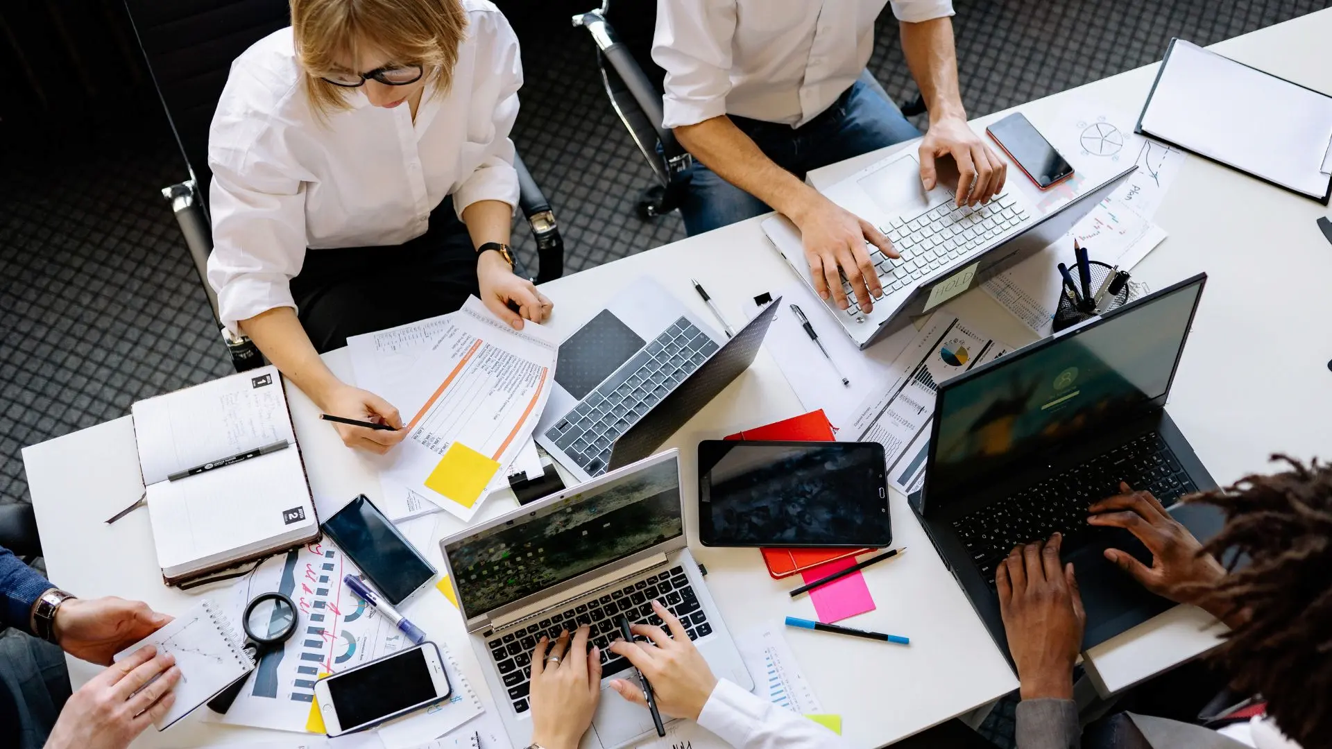 Tavolo di lavoro con più persone impegnate su computer portatili, documenti stampati, taccuini e grafici, a rappresentare attività di analisi, gestione amministrativa e organizzazione del lavoro.