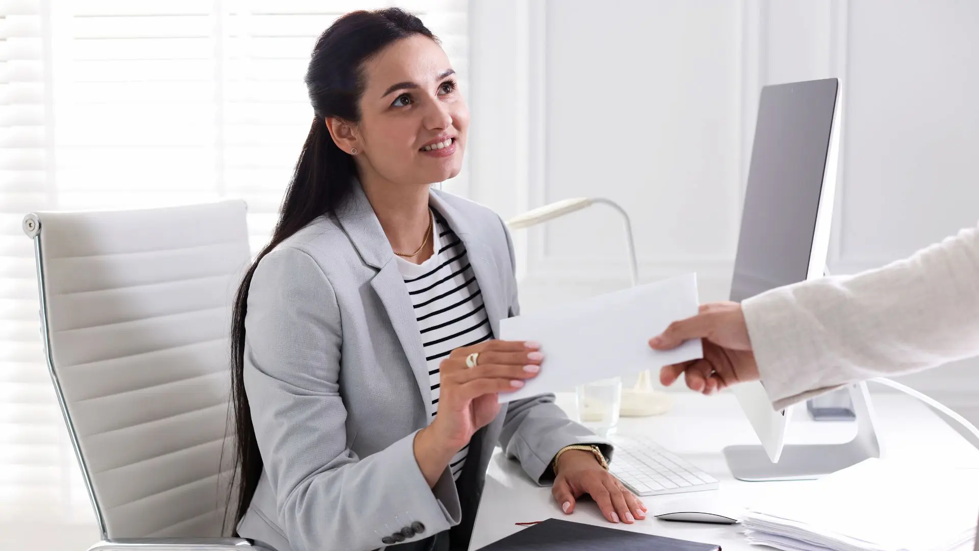 Persona seduta a una scrivania in un ufficio moderno mentre riceve una busta da un’altra persona, con documenti e computer sul tavolo, a rappresentare procedure amministrative legate alle assunzioni o alla gestione di incentivi occupazionali.