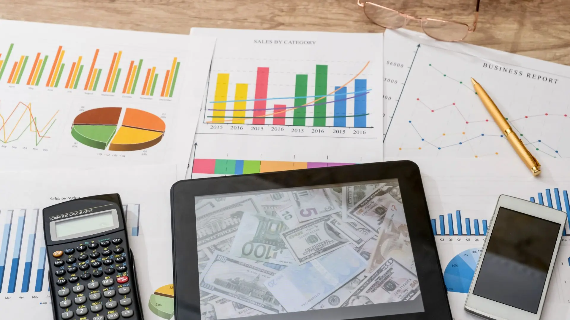 Scrivania con fogli che riportano grafici e report aziendali, una calcolatrice, uno smartphone, una penna e un tablet su cui sono visibili banconote, a rappresentare il calcolo del ticket di licenziamento e gli oneri contributivi a carico del datore di lavoro.