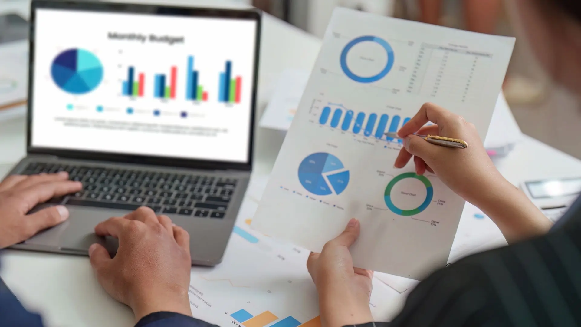 Scrivania con computer portatile che mostra grafici, fogli con diagrammi e una persona che tiene un documento con statistiche, a rappresentare analisi di budget e gestione dei fringe benefit.