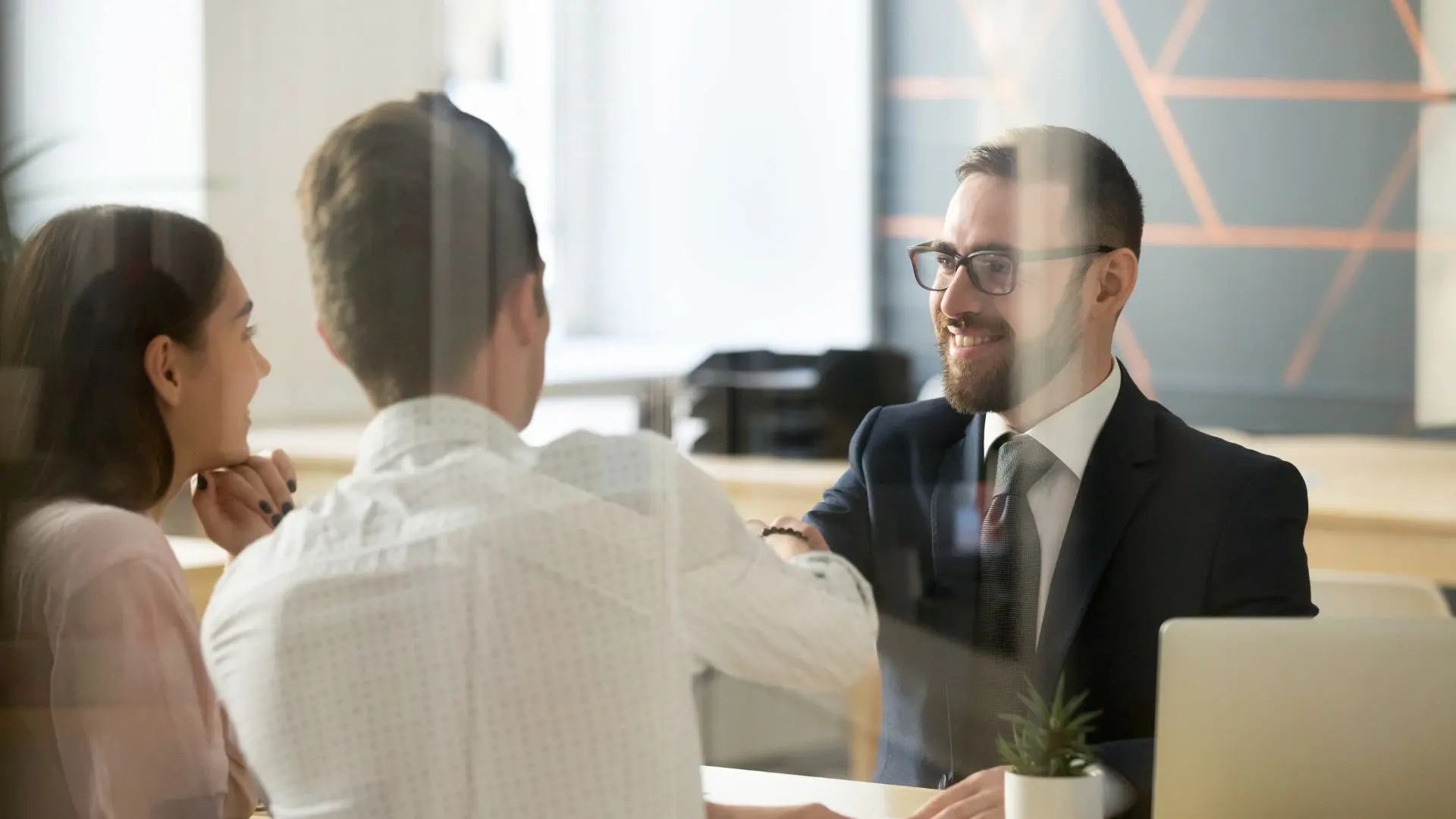 Tre persone sedute attorno a un tavolo in un ufficio moderno, durante un incontro professionale o colloquio, mentre due di loro si stringono la mano, a rappresentare orientamento e accompagnamento al lavoro.