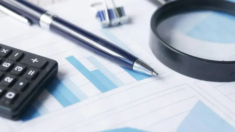 Scrivania con documenti contenenti grafici, una calcolatrice, una penna e una lente d’ingrandimento appoggiati sopra, a rappresentare analisi dei massimali e verifiche sugli aiuti De Minimis.