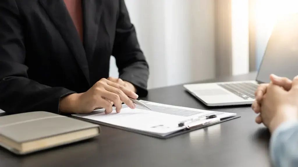 Persona seduta a un tavolo che illustra un documento con una penna a un’altra persona, in un ambiente d’ufficio, con un laptop e un blocco appunti sul tavolo, a rappresentare attività di consulenza e verifica della documentazione per il modello OT23.