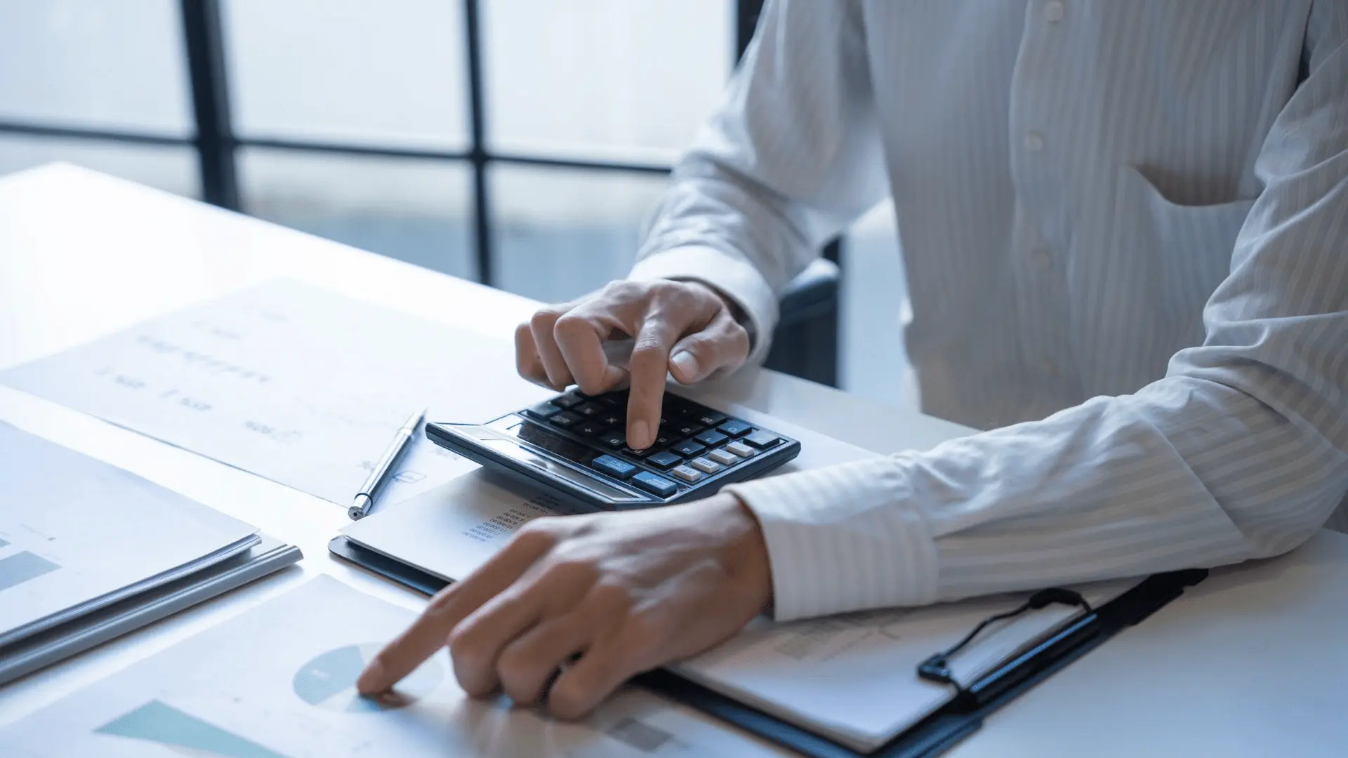 Persona che utilizza una calcolatrice e indica dati su documenti con grafici, su una scrivania con fogli e un computer portatile, a rappresentare calcoli fiscali e determinazione del costo del personale.