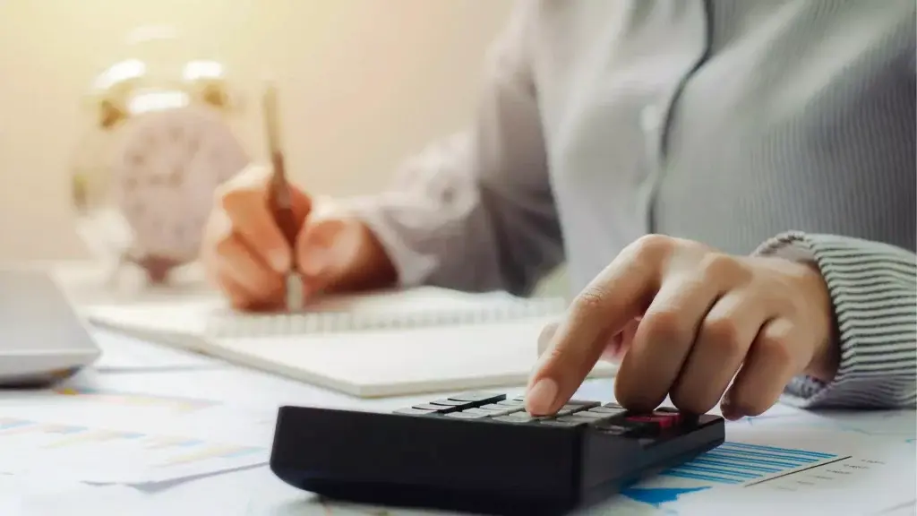 Persona alla scrivania che utilizza una calcolatrice e prende appunti con un taccuino, accanto a documenti e un computer portatile, a rappresentare attività amministrative e calcoli richiesti per la domanda di riduzione contributiva nei contratti di solidarietà.