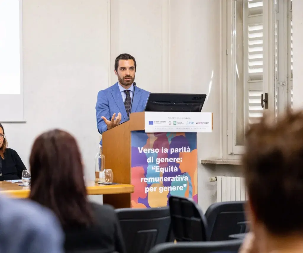 Relatore che interviene a un seminario in una sala conferenze, posizionato dietro un podio con pannello dedicato al tema “Verso la parità di genere: equità remunerativa per genere”, di fronte a un pubblico seduto.