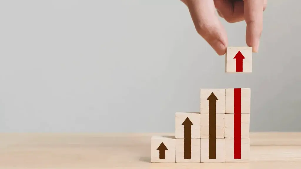 Mano che posiziona un cubo di legno con una freccia verso l’alto sopra una pila di altri cubi disposti a gradini, ciascuno con una freccia verticale stampata, a rappresentare crescita, attivazione di opportunità e progressione strategica.