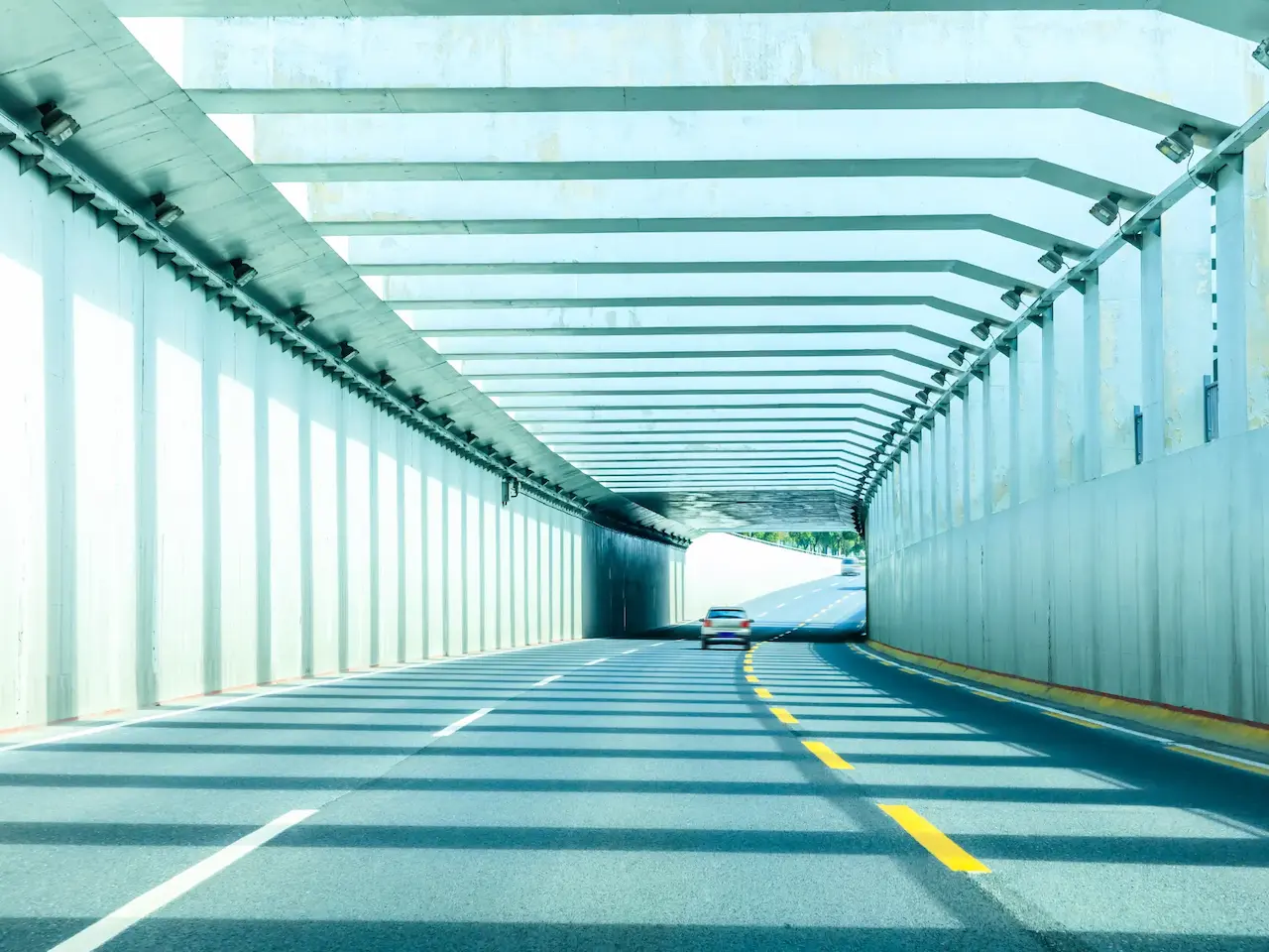 Auto in movimento all'interno di un tunnel autostradale moderno caratterizzato da un gioco di luci e ombre geometriche. Immagine rappresentativa di un'azienda capogruppo nel settore della gestione di strade, ponti e gallerie.