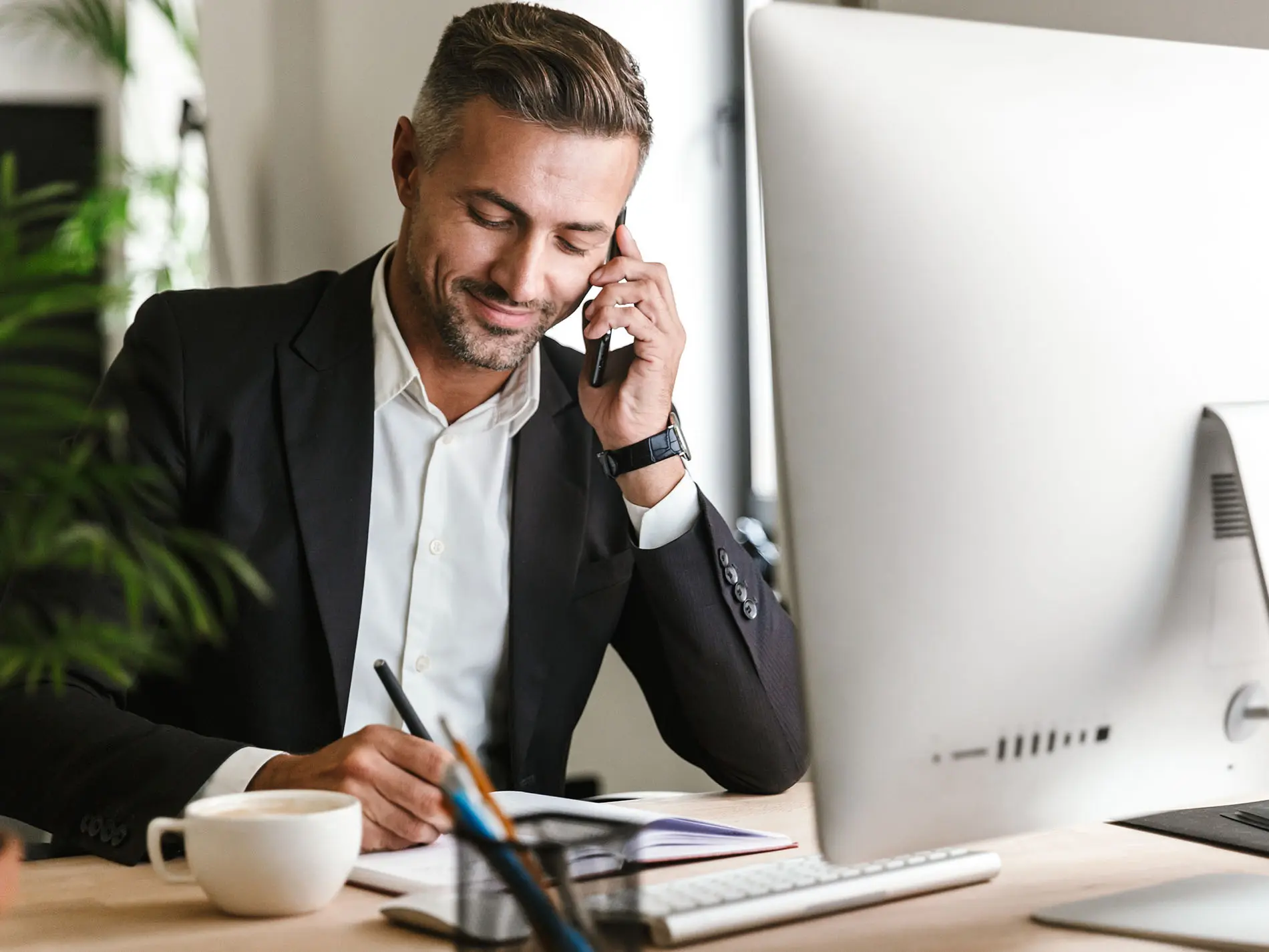Uomo d'affari sorridente che parla al cellulare e prende appunti alla sua scrivania davanti a un computer. Immagine rappresentativa per un'agenzia che offre servizi alle imprese e campagne di marketing.