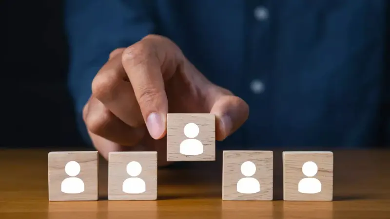 Mano che posiziona un cubo di legno con l’icona di una persona sopra altri cubi simili, simbolo di selezione delle risorse umane e politiche di assunzione
