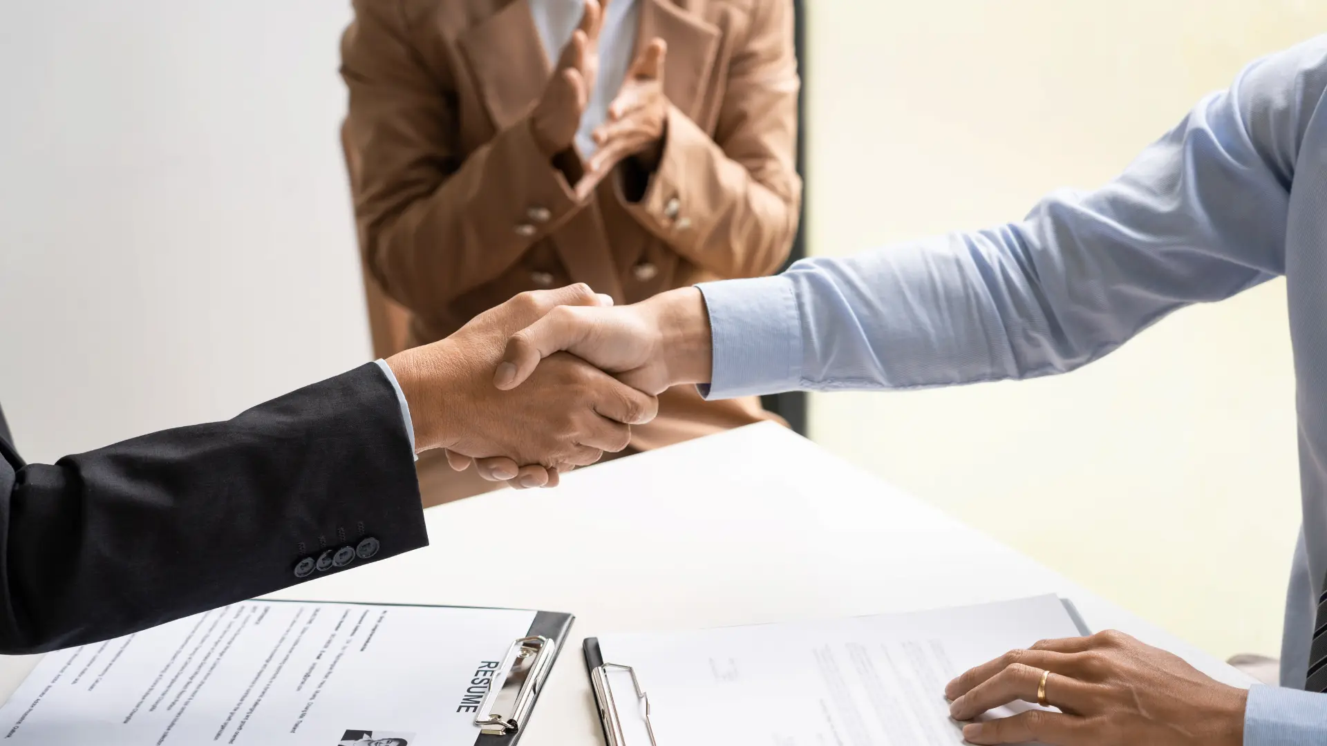 Primo piano di due persone in abiti da lavoro che si stringono la mano sopra una scrivania, con una terza persona sfocata sullo sfondo che applaude. Sulla scrivania sono visibili dei documenti, tra cui uno con la scritta 