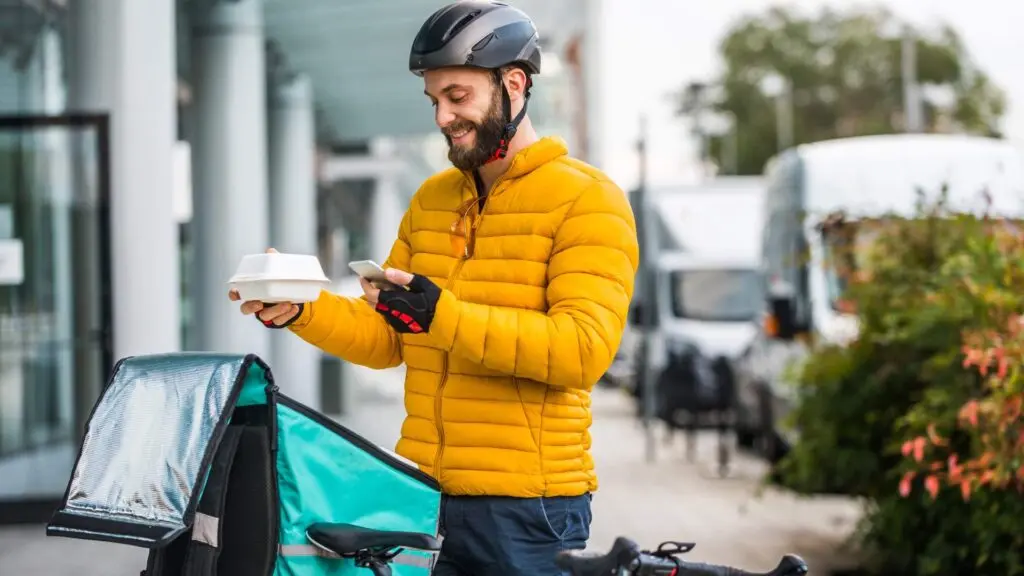 Un rider con giacca gialla e casco controlla lo smartphone accanto a una bicicletta con borsa termica per consegne a domicilio. Immagine rappresentativa dell'ottimizzazione dei costi del lavoro nel settore Food Delivery.