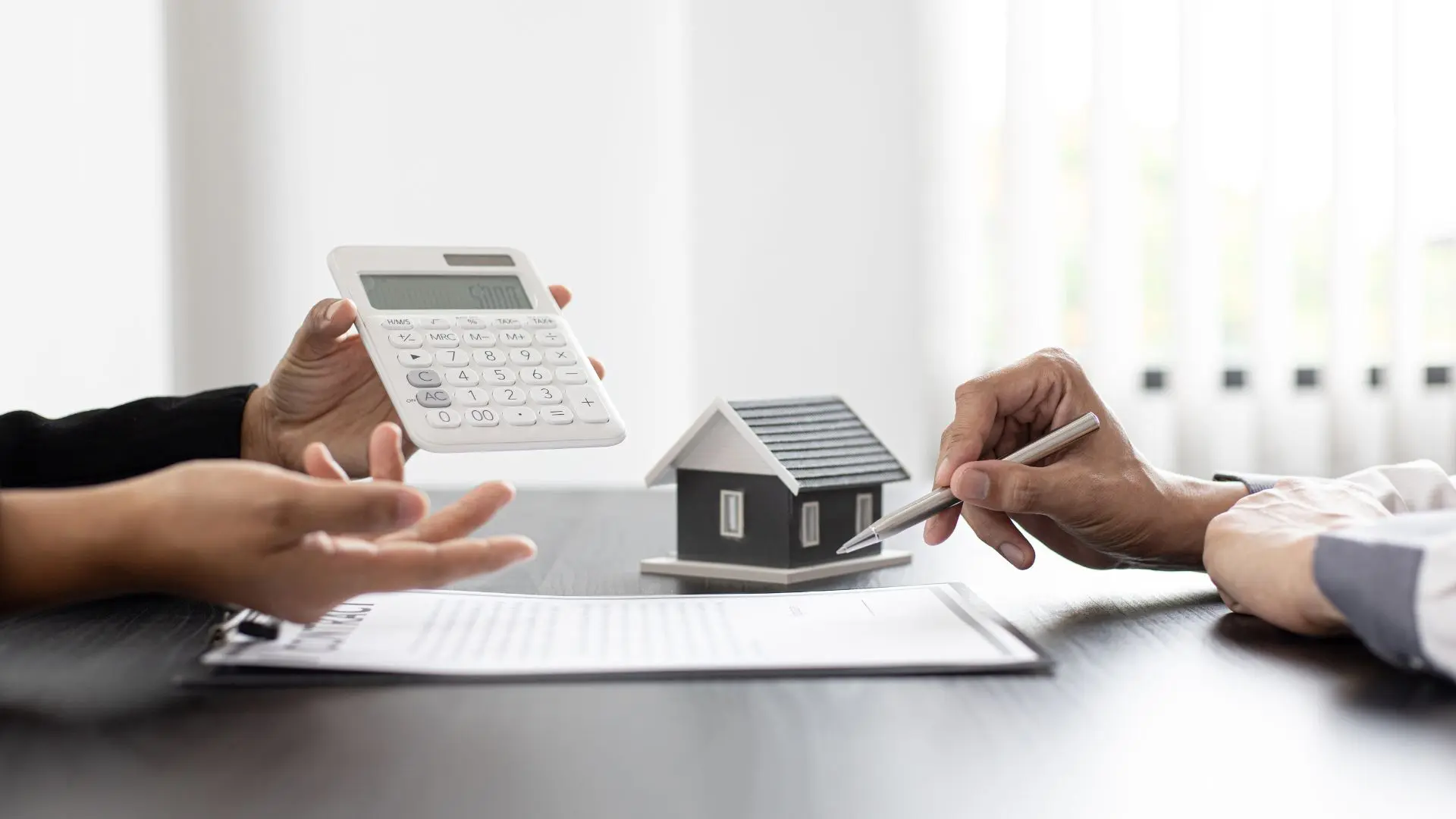 Primo piano di due persone che analizzano un contratto con una calcolatrice e un modello di casa su un tavolo da ufficio.