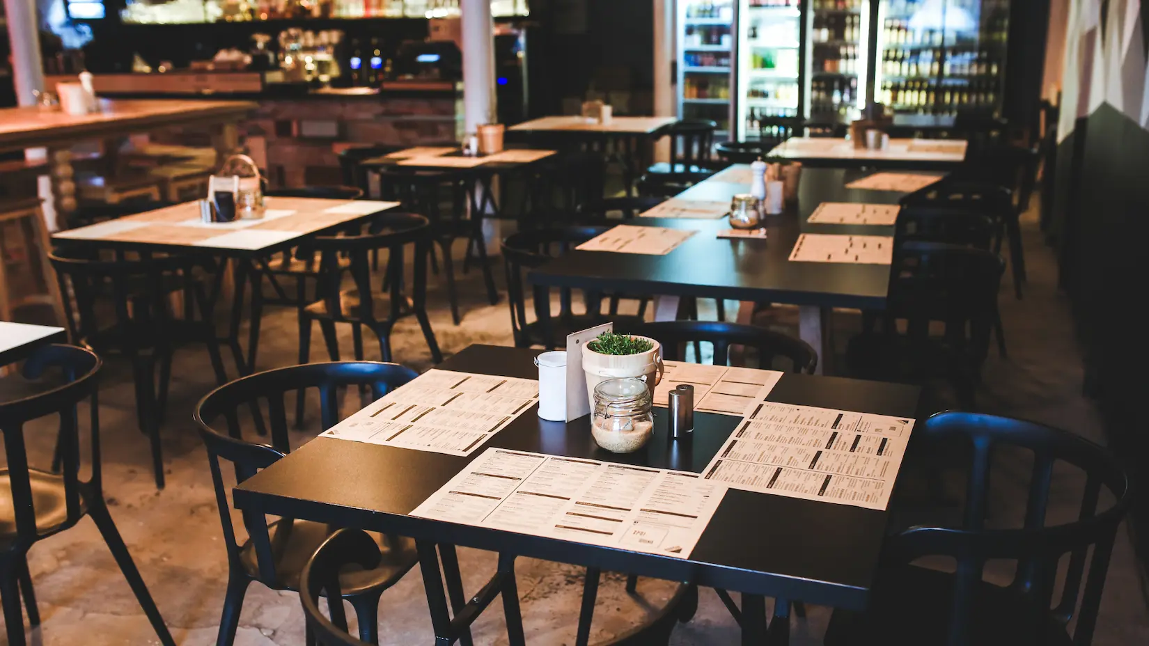 Sala di un ristorante con tavoli neri apparecchiati con tovagliette menu, sedie scure e bancone bar sullo sfondo